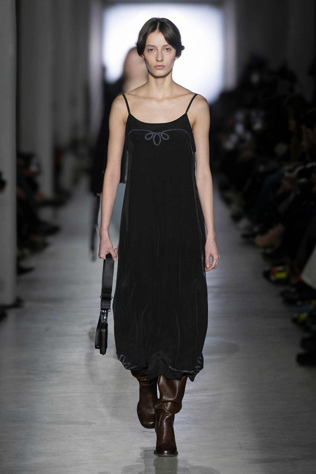 Model walks the runway in a black spaghetti-strap midi dress with a subtle stitched pattern, brown knee-high boots, and a small black bag.