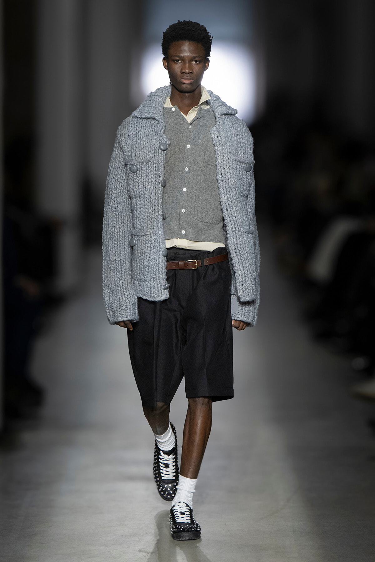 Male model on a runway wearing a chunky gray cardigan over a gray vest and beige shirt with black knee-length shorts and studded shoes.