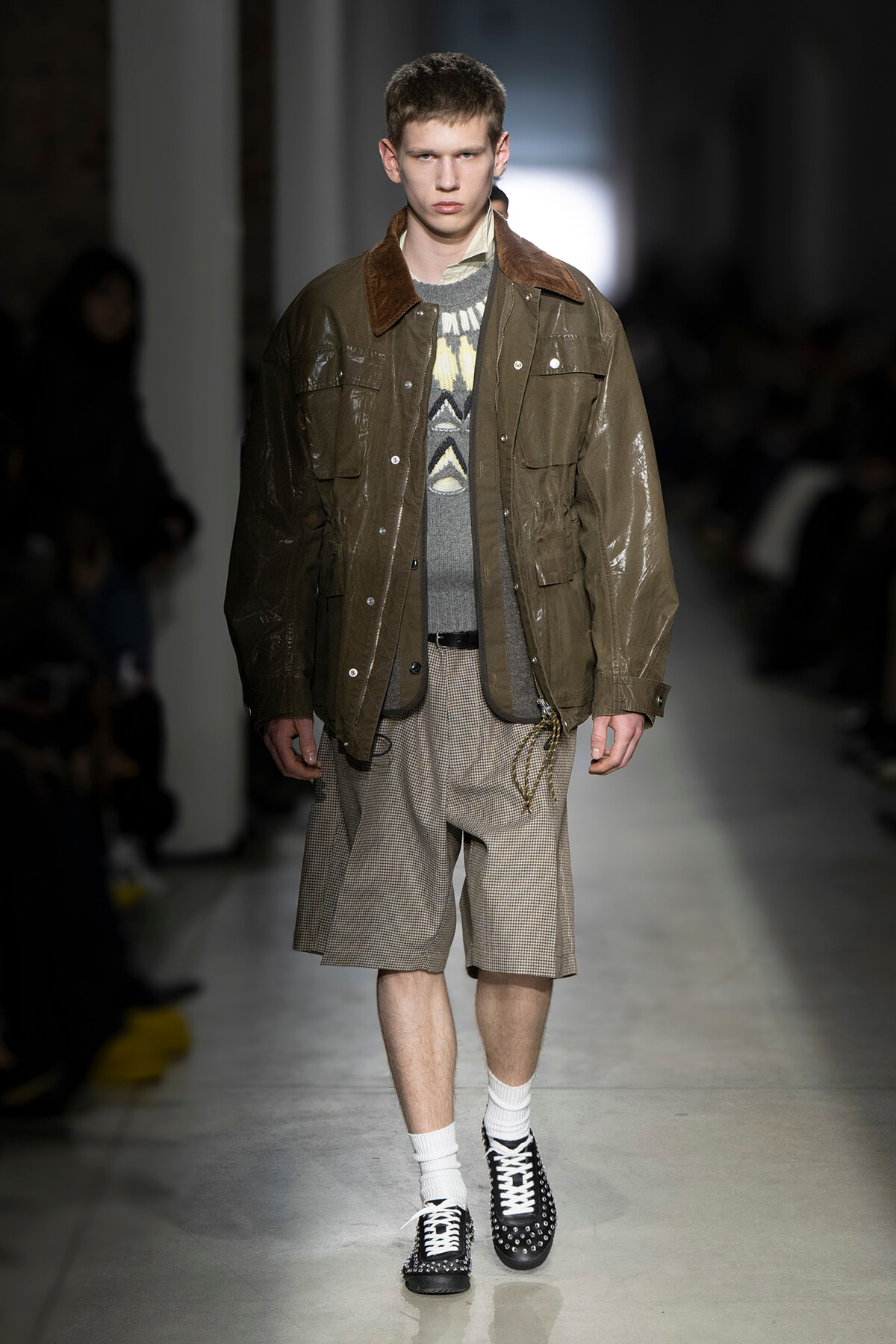 Male model walks the runway in a brown waxed jacket with a shearling collar over a patterned knit sweater and checkered shorts, black studded shoes.