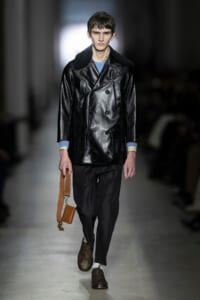Male model walks a runway wearing a black leather double-breasted coat with a shearling collar, gray pants, and brown brogues, carrying a tan pouch with a strap.