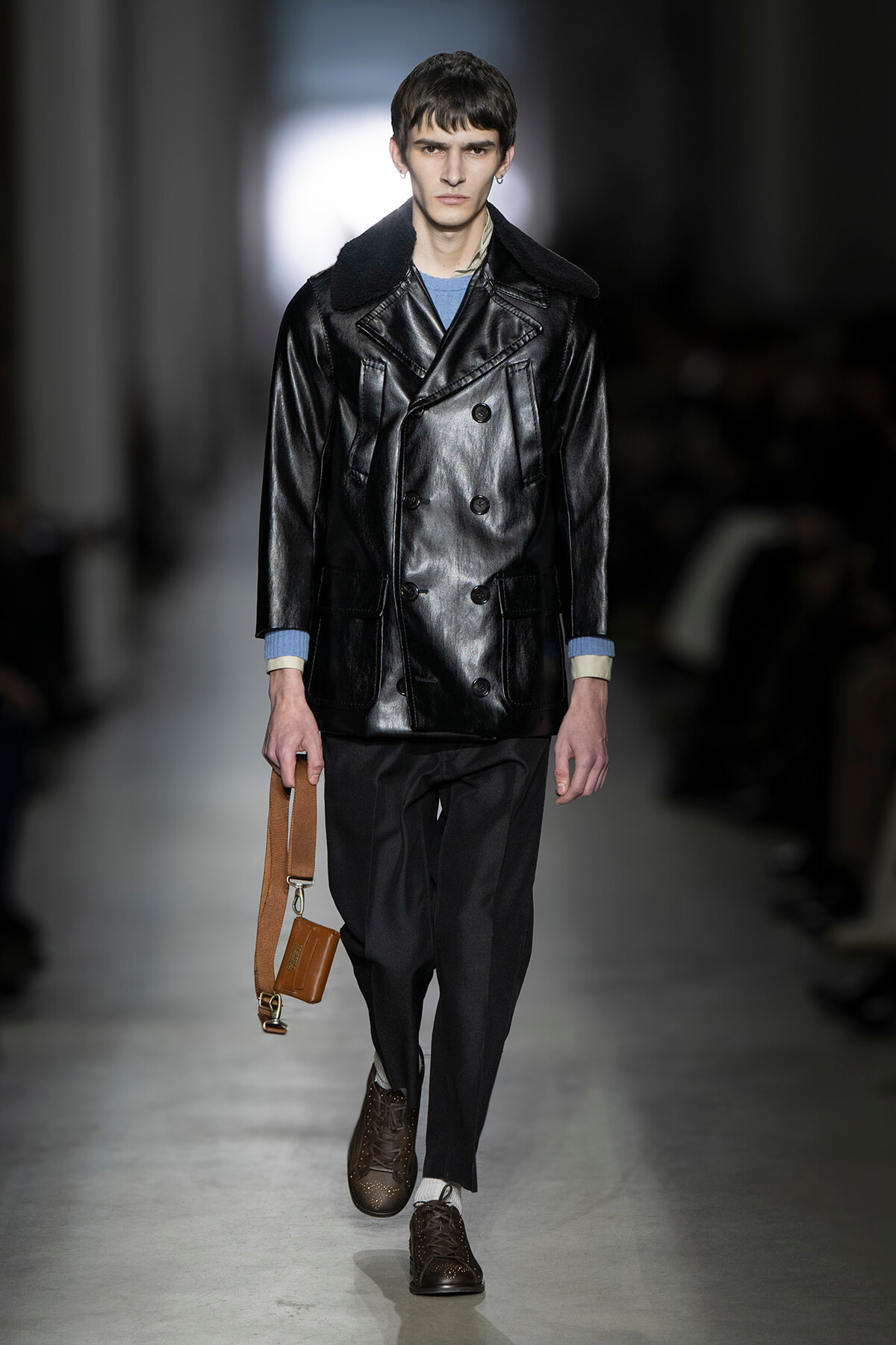Male model walks a runway wearing a black leather double-breasted coat with a shearling collar, gray pants, and brown brogues, carrying a tan pouch with a strap.