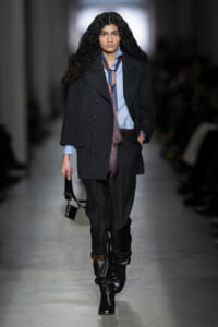 Model walking the runway in an oversized dark coat over a light blue shirt, scarf, black trousers, tall boots, and a small black bag.