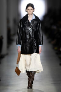 Model walking a runway in a black leather double-breasted coat, light blue shirt, cream skirt, brown boots, and a brown handbag.