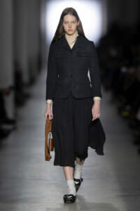 Model on a runway wearing a dark tailored jacket, flowing black skirt, white socks, black shoes, and a brown handbag nearby.
