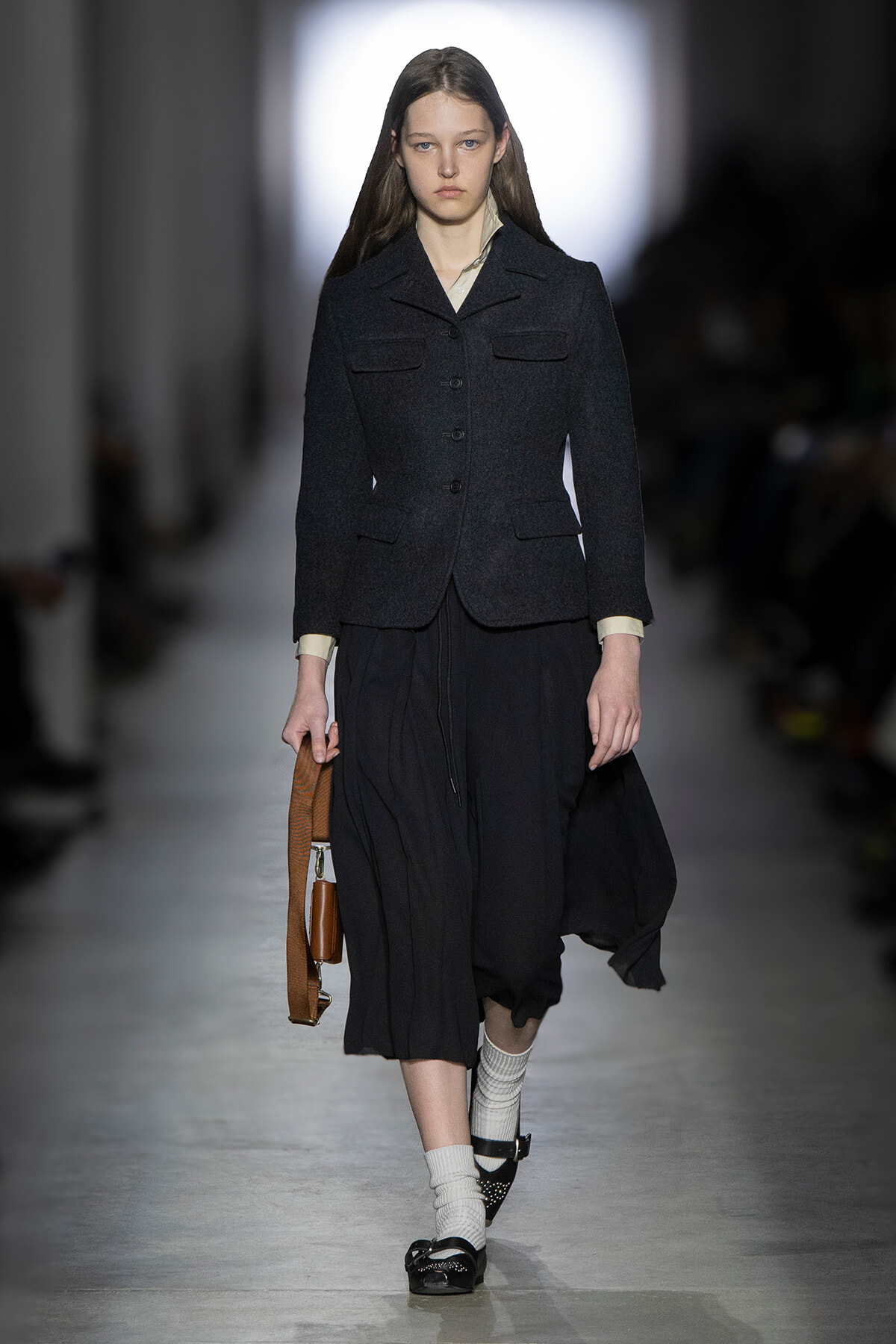 Model on a runway wearing a dark tailored jacket, flowing black skirt, white socks, black shoes, and a brown handbag nearby.