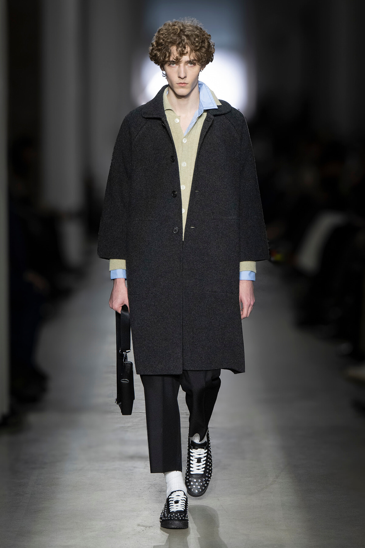 Male model walks a runway in a dark wool overcoat over a beige cardigan and light blue shirt, black trousers, studded black-and-white shoes, carrying a black bag.
