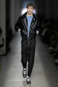 Male model walking the runway in a black leather bomber jacket, light blue sweater, black trousers, and studded sneakers.