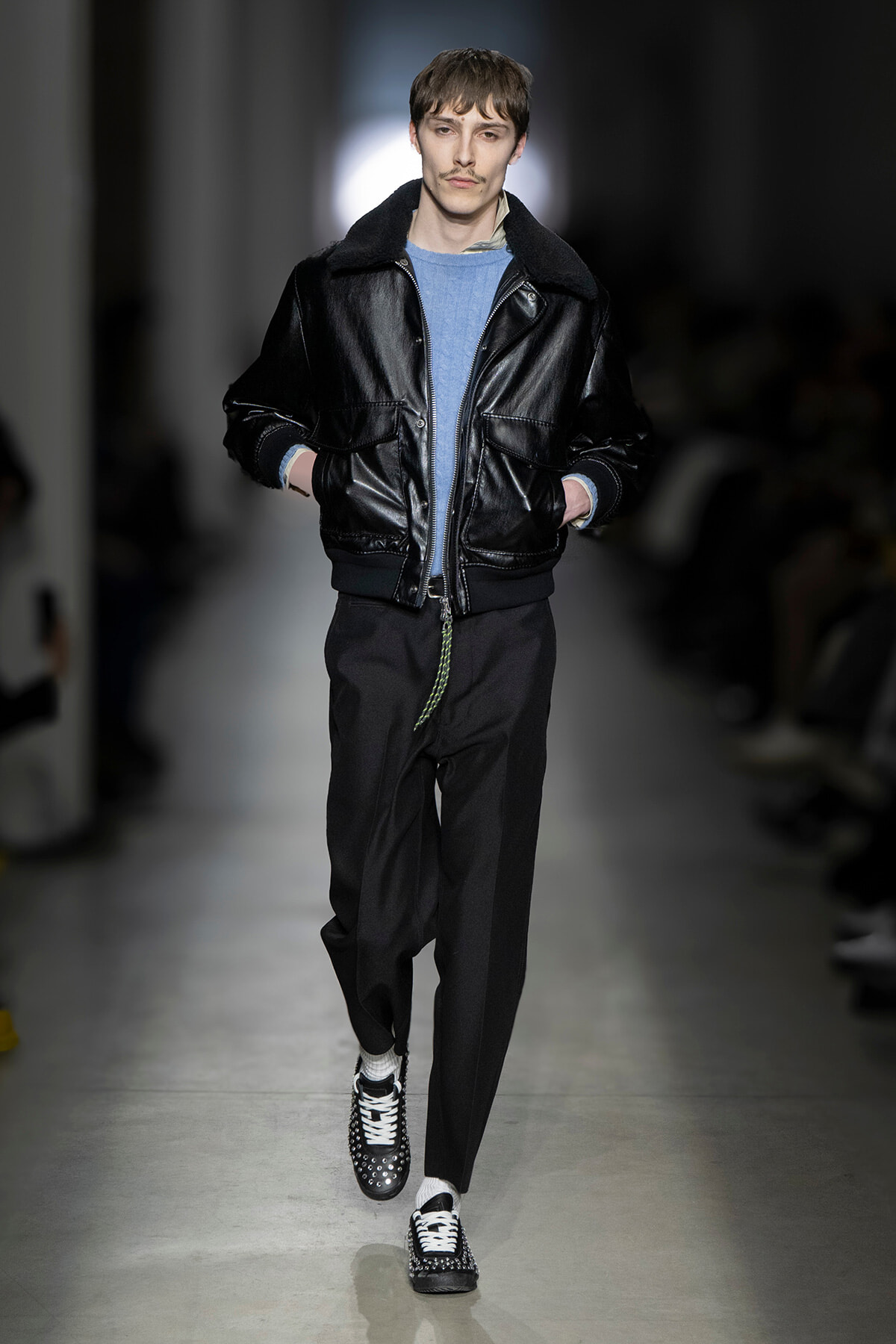 Male model walking the runway in a black leather bomber jacket, light blue sweater, black trousers, and studded sneakers.