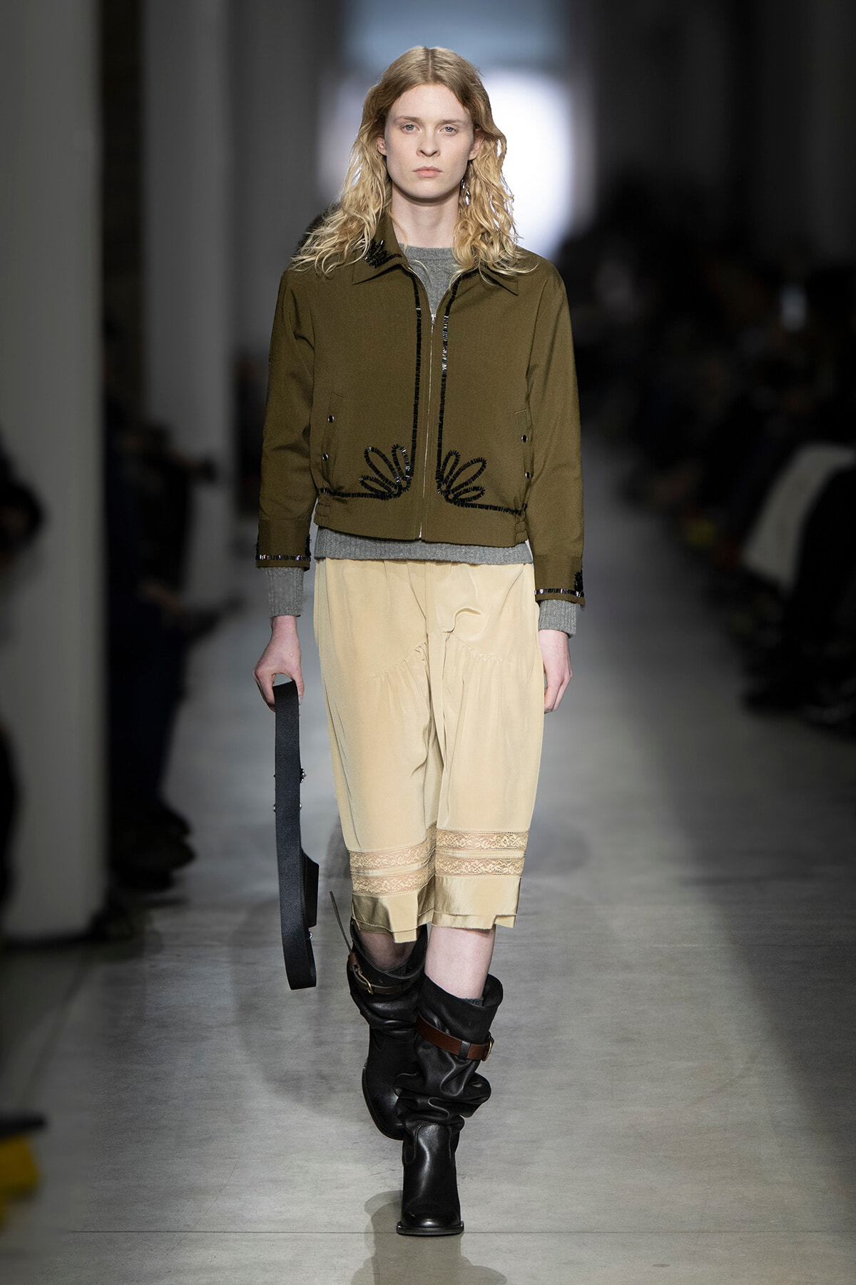 Model walks the runway in an olive cropped jacket with black embroidery, paired with beige knee-length pants with lace trim and slouchy black boots, holding a strap.
