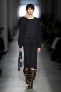 Model walking a runway in all-black ensemble: long-sleeve top with puff shoulders, wide-leg pants, brown slouch boots, and a strap bag hanging at her side