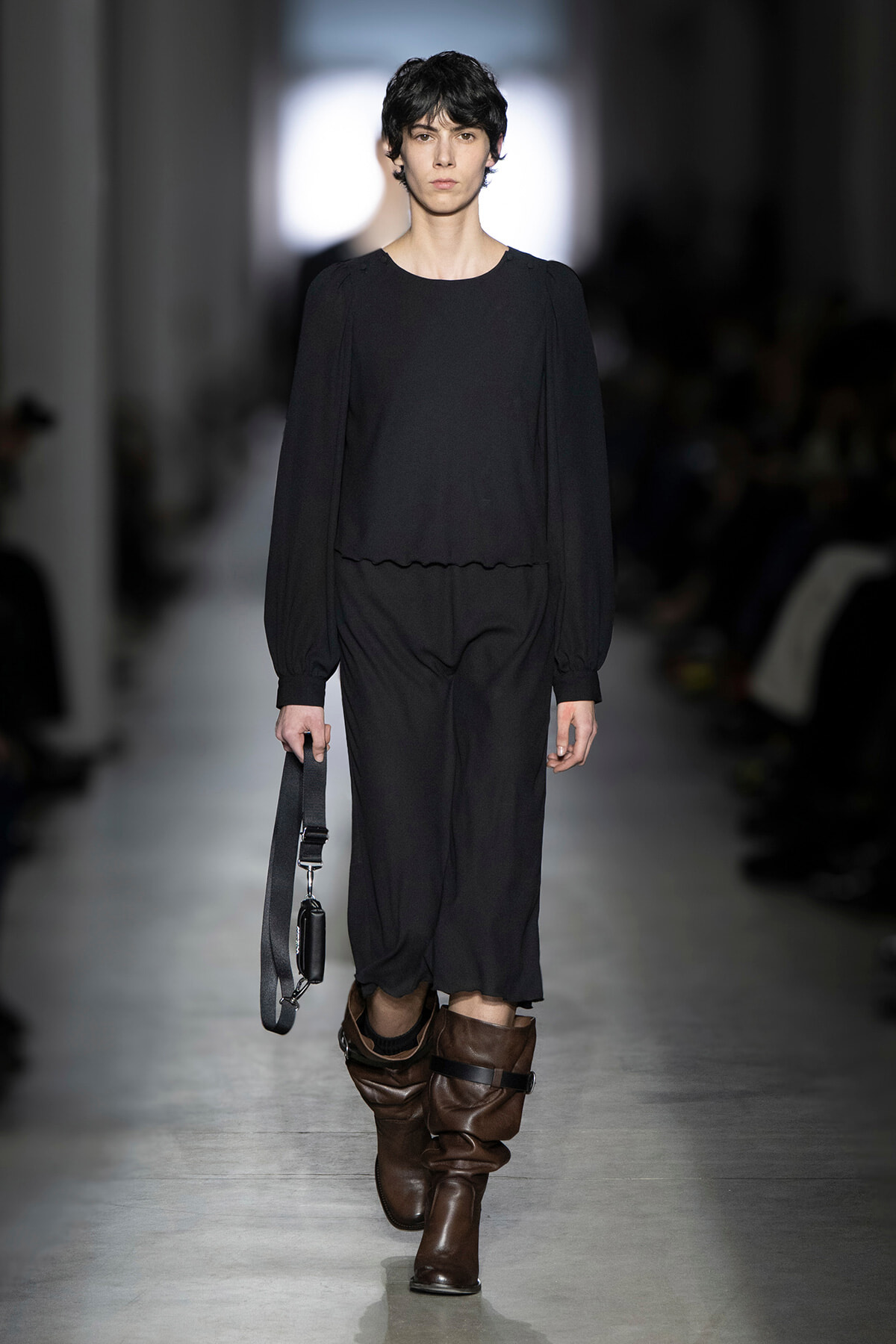 Model walking a runway in all-black ensemble: long-sleeve top with puff shoulders, wide-leg pants, brown slouch boots, and a strap bag hanging at her side