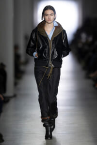 Model walks the runway in a black leather bomber jacket over a light blue shirt, dark trousers, and black boots.