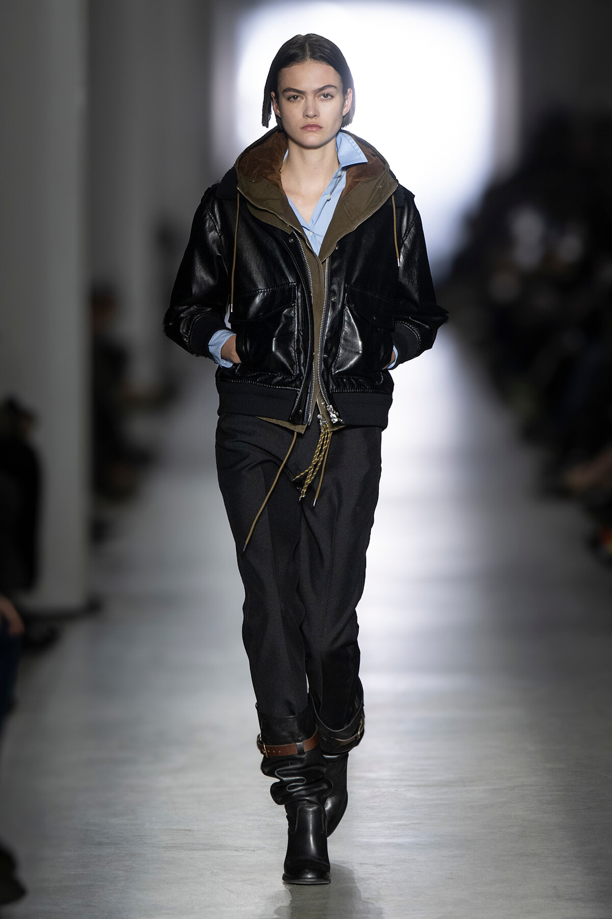 Model walks the runway in a black leather bomber jacket over a light blue shirt, dark trousers, and black boots.