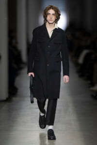 Male model walking a fashion runway in a knee-length black coat with large front pockets, white shirt, black trousers, and studded black shoes, carrying a black bag.