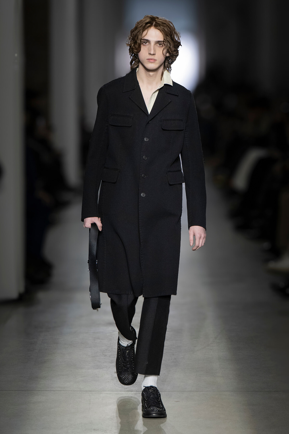 Male model walking a fashion runway in a knee-length black coat with large front pockets, white shirt, black trousers, and studded black shoes, carrying a black bag.