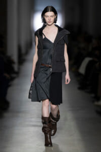 Model walks a runway in a dark layered outfit: charcoal sleeveless vest over a gray dress with a brown belt and knee-high brown boots.