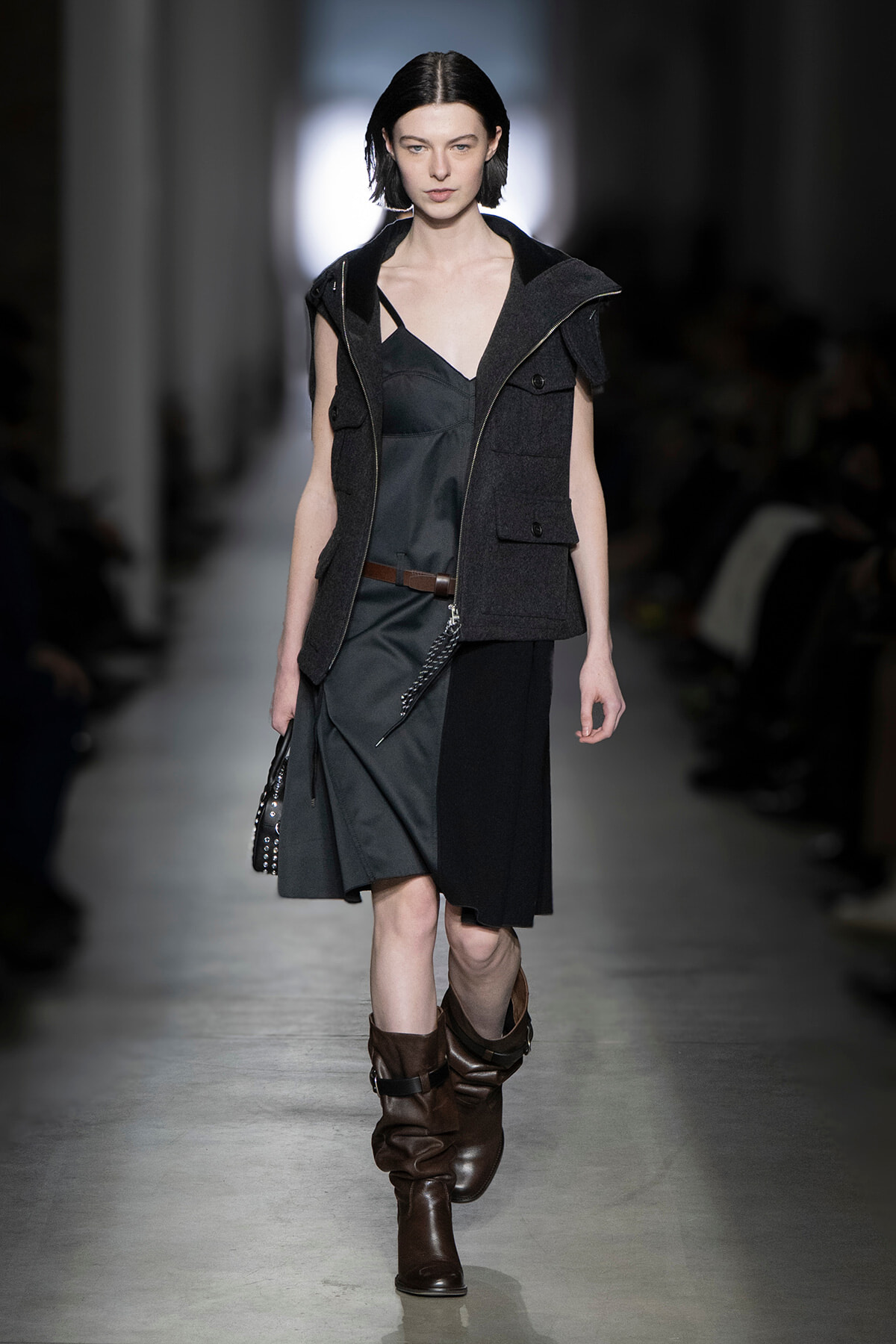 Model walks a runway in a dark layered outfit: charcoal sleeveless vest over a gray dress with a brown belt and knee-high brown boots.