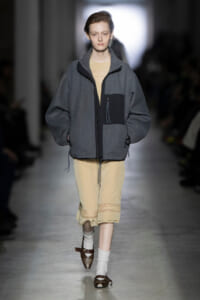 Runway model in a loose gray zip jacket over a beige dress with lace hem, walking down a fashion show runway.