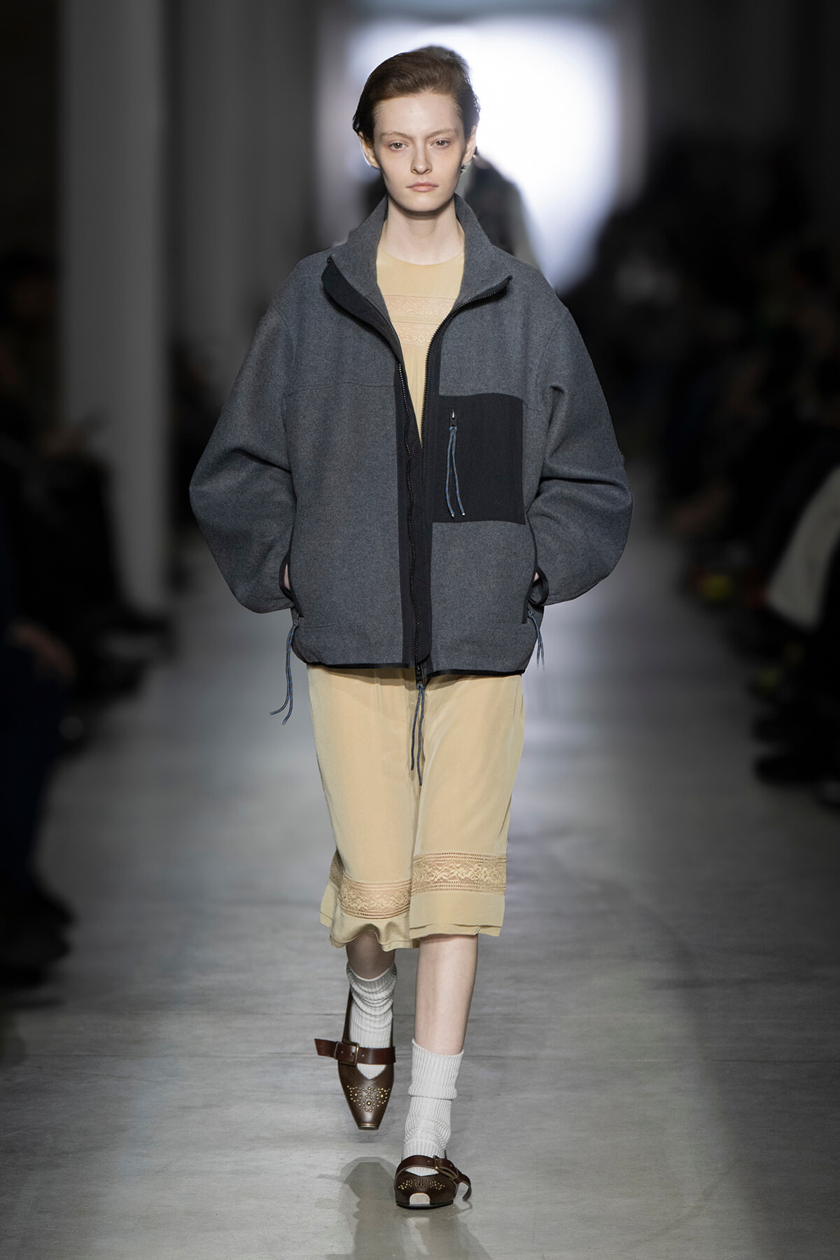 Runway model in a loose gray zip jacket over a beige dress with lace hem, walking down a fashion show runway.