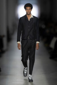 Male model walks a fashion runway wearing a black three‑button blazer, black trousers, cream shirt cuffs, and studded black-and-white shoes.