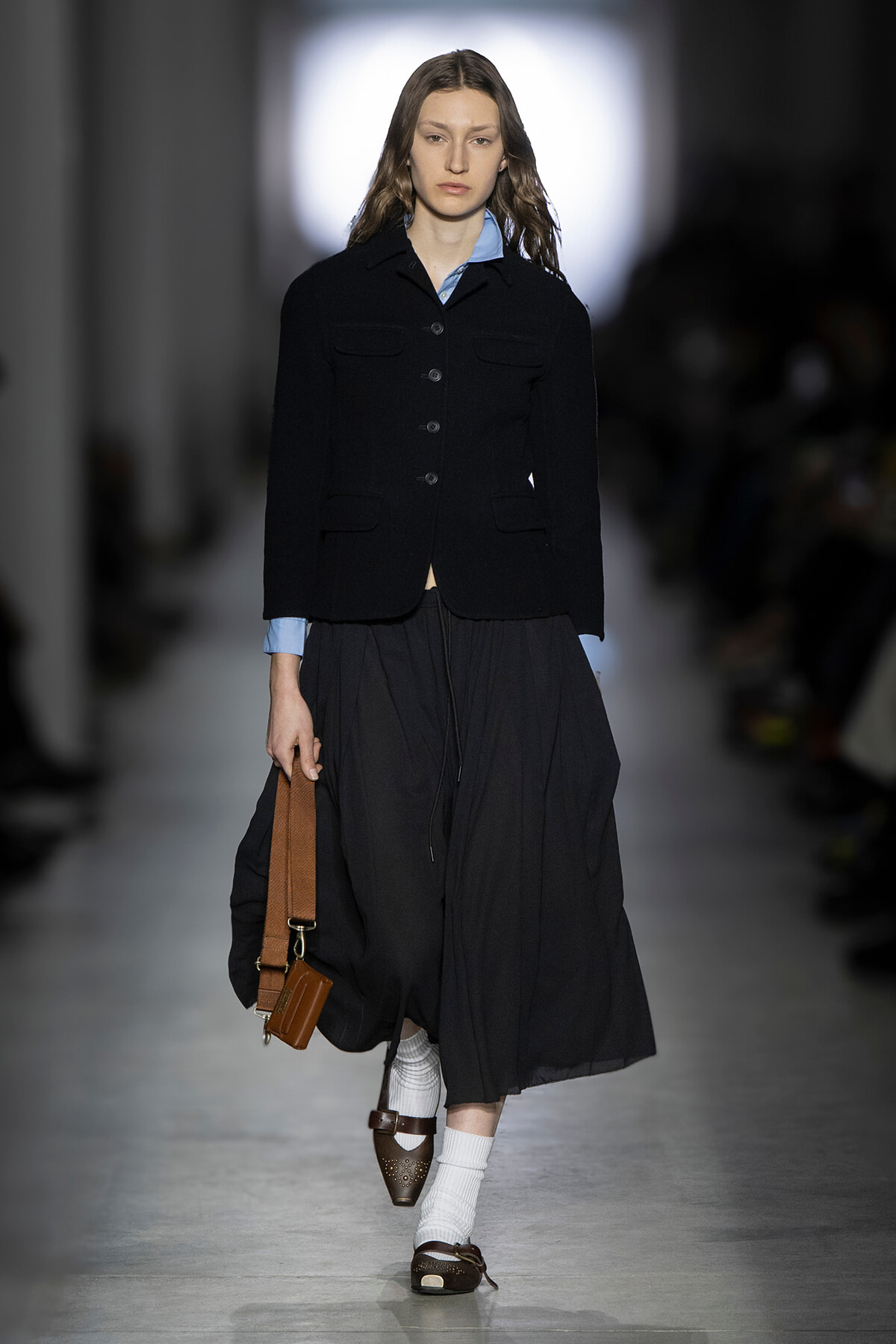 Model walking the runway in a black cropped blazer over a light blue shirt, paired with a long black skirt and brown accessories.
