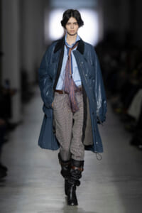 Female model walking a runway in a blue oversized coat over a light blue shirt, with a long fringe necktie, plaid pants, and tall black boots at a fashion show.