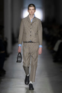 Male runway model wearing a beige checked suit with blue shirt cuffs, carrying a small black studded handbag on the catwalk