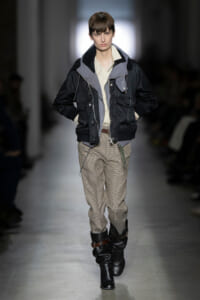 Runway model wearing a layered black jacket over a light shirt, plaid trousers, and tall black boots on a fashion catwalk.