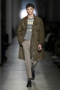 Male model walking runway in an oversized olive-brown raincoat over a patterned gray sweater and checkered trousers; black shoes complete the look.