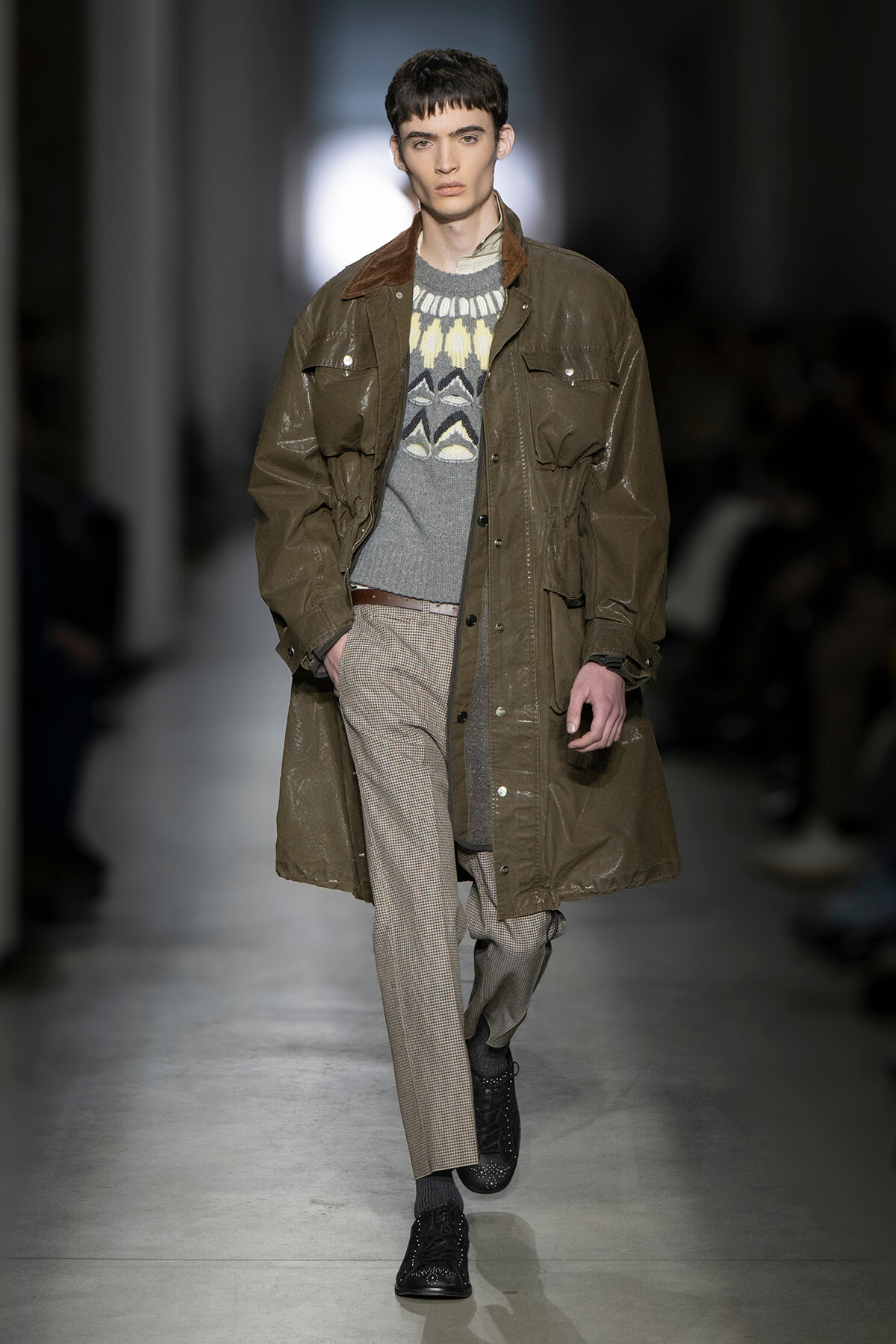 Male model walking runway in an oversized olive-brown raincoat over a patterned gray sweater and checkered trousers; black shoes complete the look.