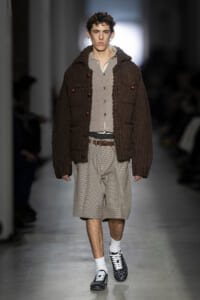 Male model on a runway wearing a bulky brown cardigan over a beige cardigan and plaid shorts with white socks and studded shoes.
