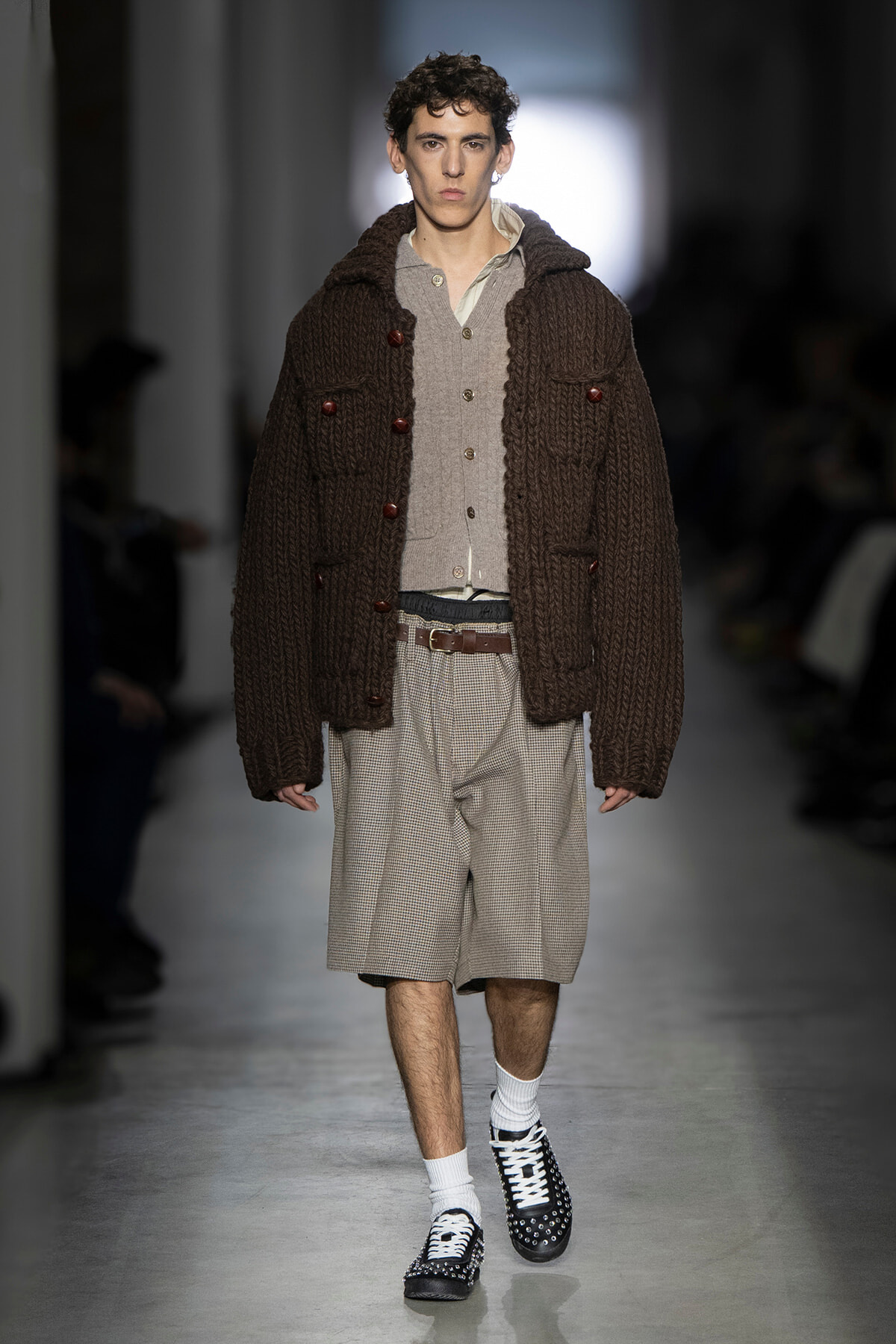 Male model on a runway wearing a bulky brown cardigan over a beige cardigan and plaid shorts with white socks and studded shoes.
