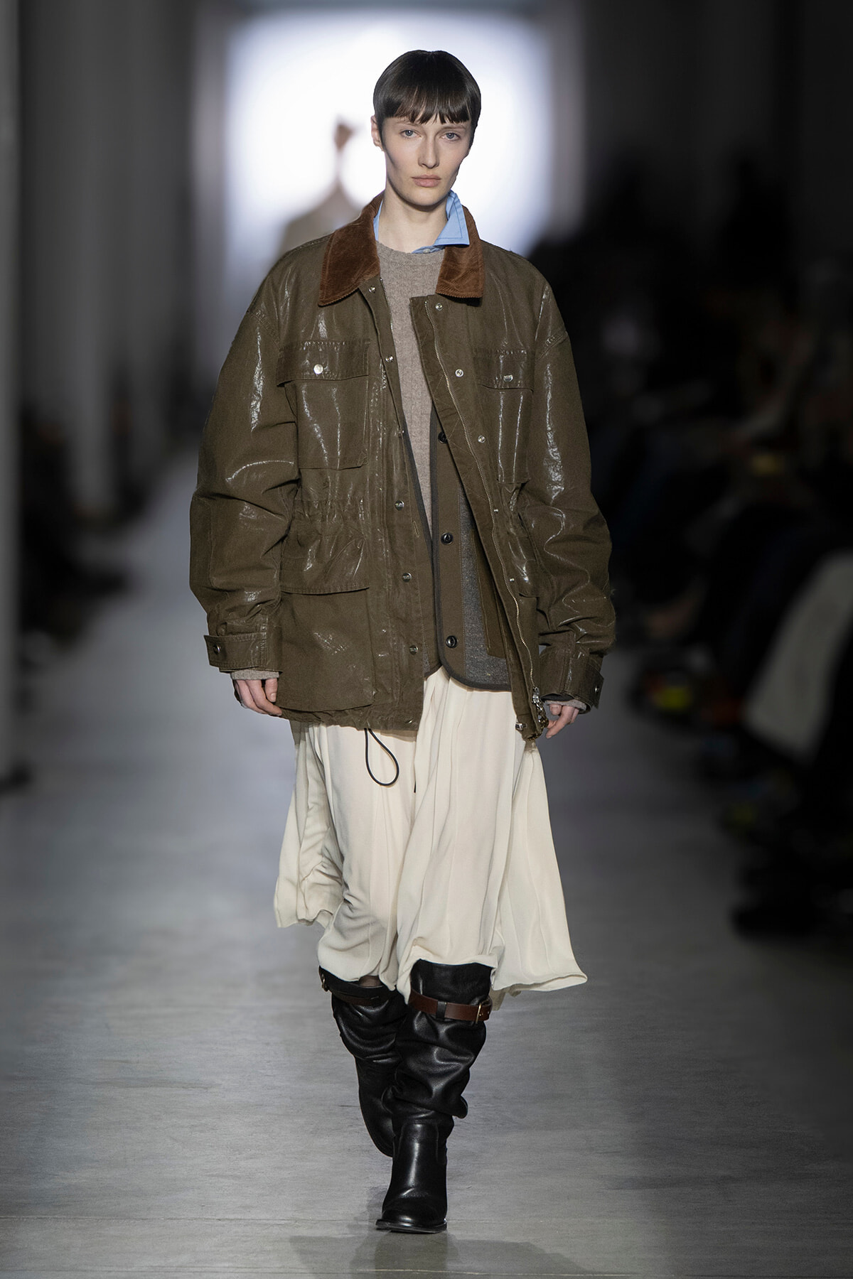 Model walks the runway in an oversized olive parka with brown collar, layered over neutral knits and a cream flowing skirt paired with tall black boots.