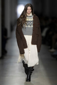 A model walks a fashion runway wearing a chunky brown knit cardigan over a gray patterned sweater and a cream flowing skirt, paired with black slouch boots.