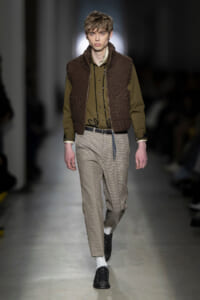 Male model walking runway in olive shirt, brown knitted vest, and beige checked trousers with black shoes and white socks, belt visible.
