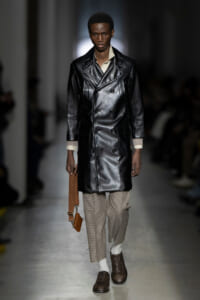 Male model on a runway wearing a black leather double-breasted coat with beige checked trousers and brown lace-up shoes, carrying a brown crossbody bag.