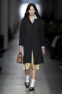 Model walking runway in a dark wool long coat over a light blue shirt, beige layered skirt, white socks, black shoes, carrying a small brown studded handbag