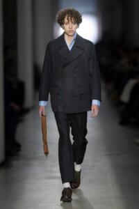 Male model on a runway wearing a dark double-breasted coat over a light blue shirt, dark trousers, white socks, brown shoes, and carrying a brown strap bag.