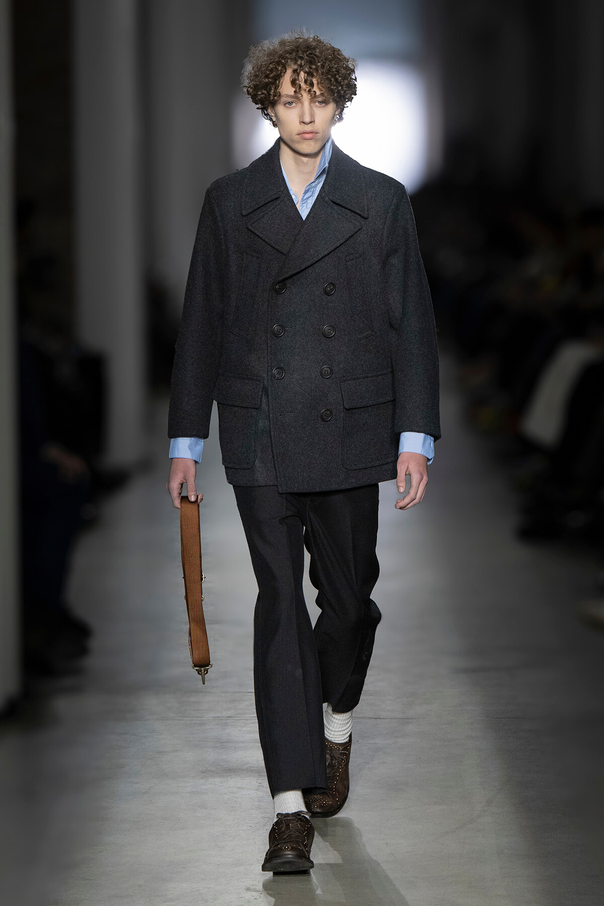 Male model on a runway wearing a dark double-breasted coat over a light blue shirt, dark trousers, white socks, brown shoes, and carrying a brown strap bag.