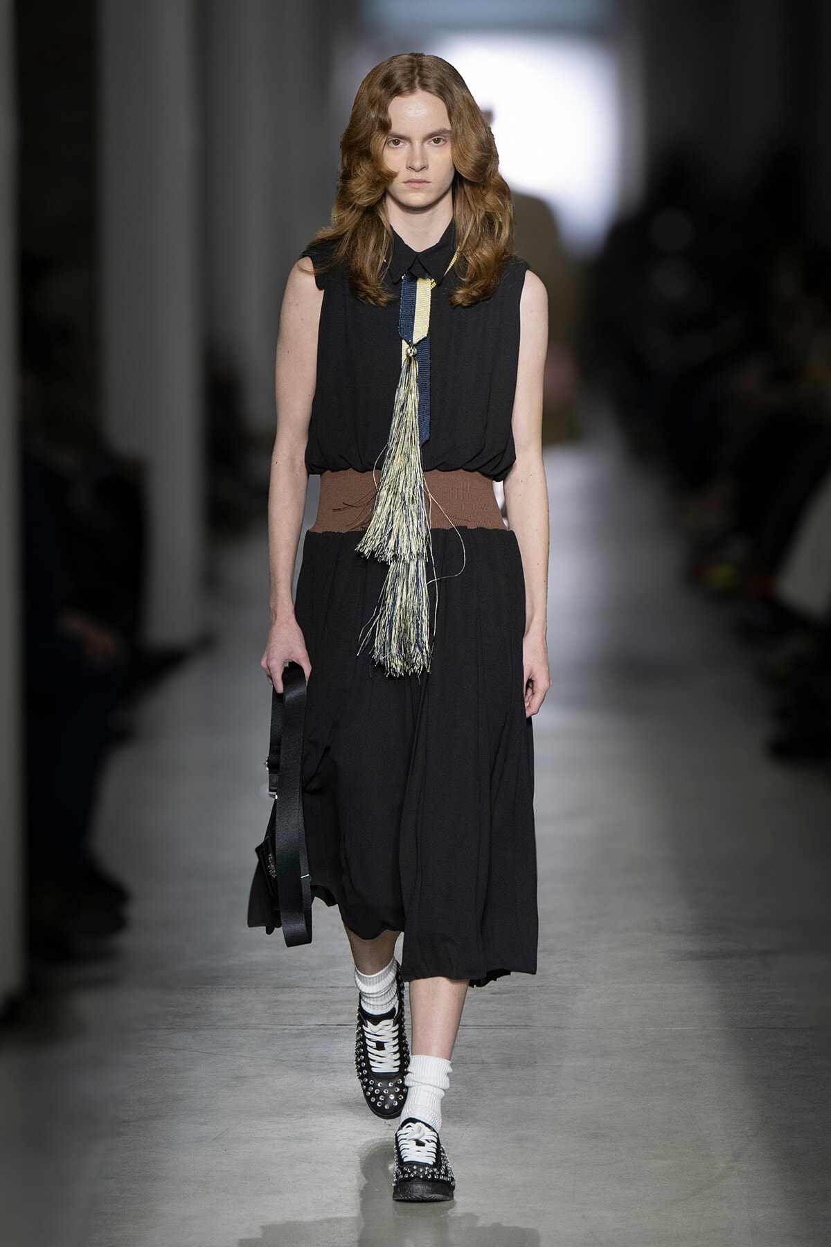 Model walks a fashion show wearing a sleeveless black jumpsuit-like dress with a wide brown belt and a long tassel necklace, black-and-white shoes, and white socks, carrying a black bag on the left.