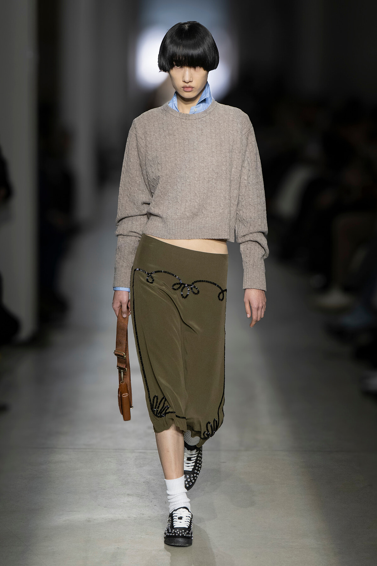 Model on a runway wearing a cropped gray knit sweater over a blue shirt, olive embroidered skirt, white socks, studded black-and-white shoes, and a brown handbag.