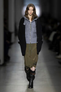 Model walking a runway in a gray zip-front utility vest over a black oversized sweater, paired with olive cargo shorts and tall black boots.