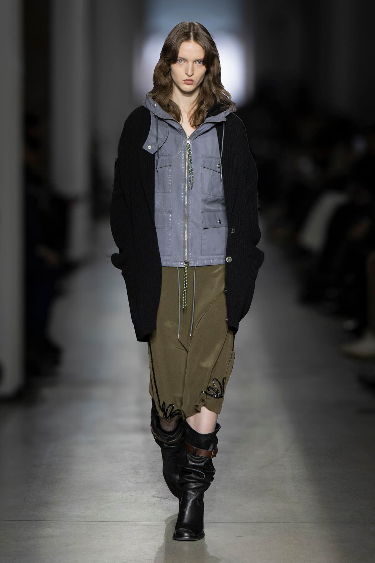 Model walking a runway in a gray zip-front utility vest over a black oversized sweater, paired with olive cargo shorts and tall black boots.