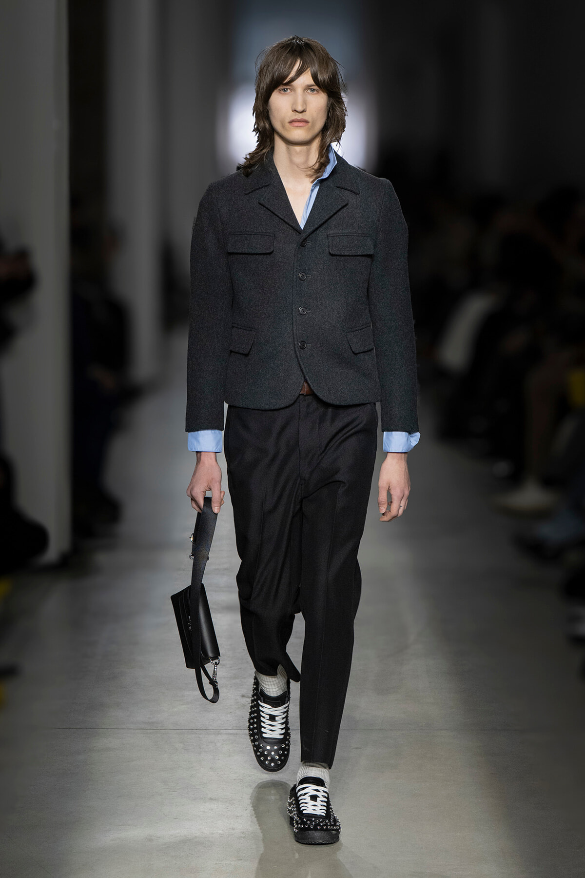 Male model on a runway wearing a dark tailored blazer over a light blue shirt, black trousers, studded black shoes, and carrying a small black bag.