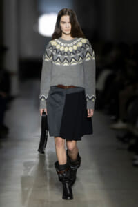 Model on a runway wearing a gray patterned knit sweater, a color-block black and gray skirt with a brown belt, slouchy black boots, and a small black handbag