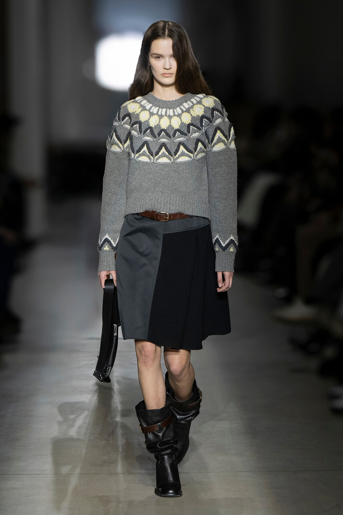 Model on a runway wearing a gray patterned knit sweater, a color-block black and gray skirt with a brown belt, slouchy black boots, and a small black handbag