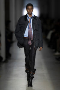 Model on a runway wearing an oversized dark jacket over a light blue shirt with a fringed, purple tie, black trousers, and slouchy black boots with brown straps.