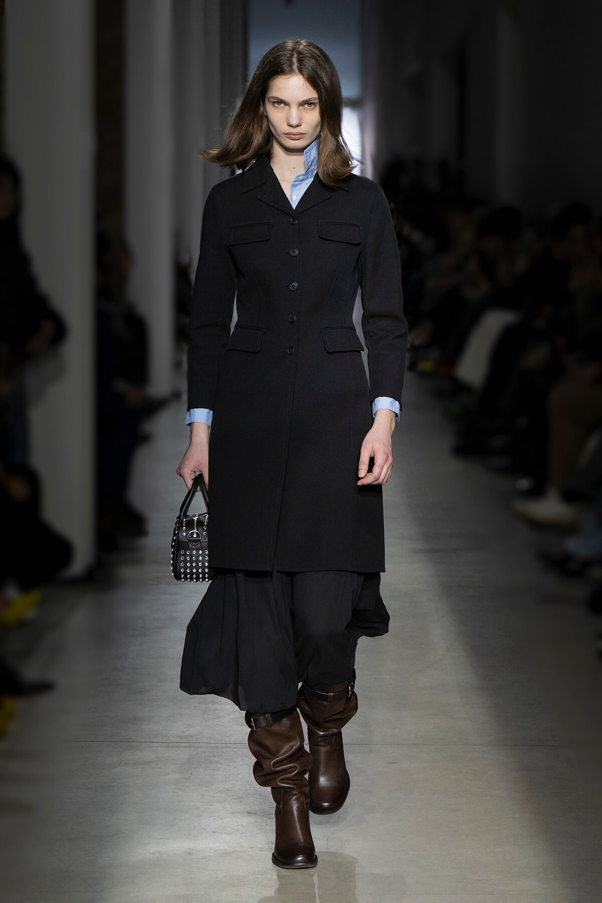 Model walking a runway in a tailored black coat over a light blue shirt, paired with a black skirt and brown knee-high boots, holding a studded handbag.