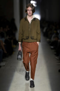 Male model on a runway wearing an olive bomber jacket with black embroidery, rust-colored trousers, and a small black studded handbag.