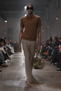 Male model on runway wearing brown knit polo, beige trousers, oversized black visor sunglasses, and carrying a metallic handbag; audience seated along the catwalk.